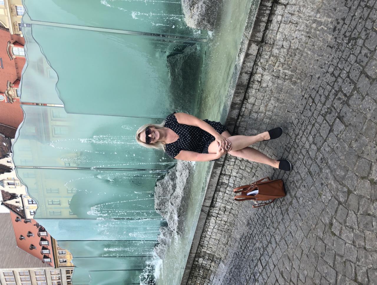 Kobieta w okularach przeciwsłonecznych siedzi przy fontannie z przezroczystego szkła na tle miejskiej zabudowy z czerwonymi dachami.