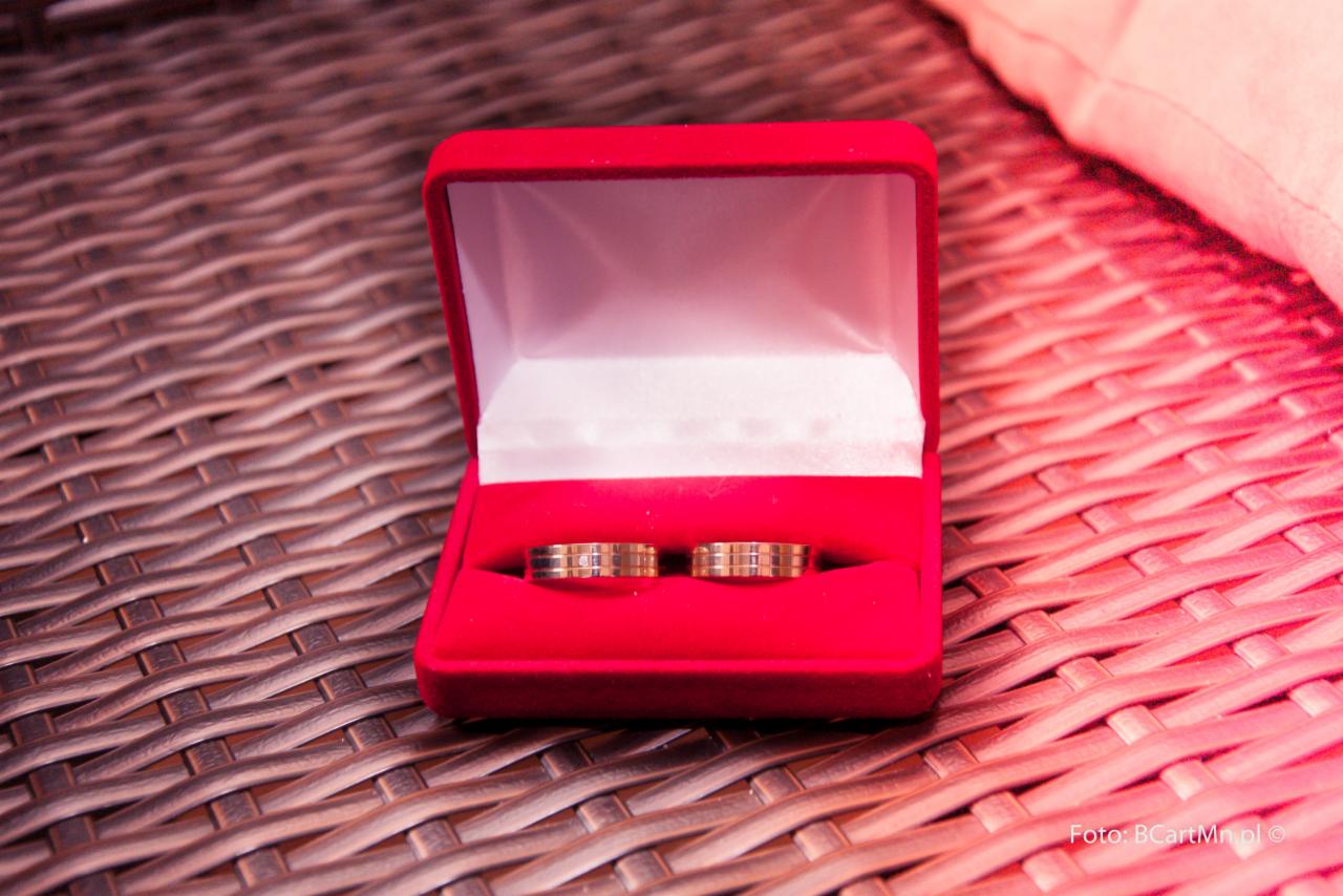 Dwa złote obrączki z delikatnym zdobieniem w czerwonym, aksamitnym pudełku na tle plecionego materiału.