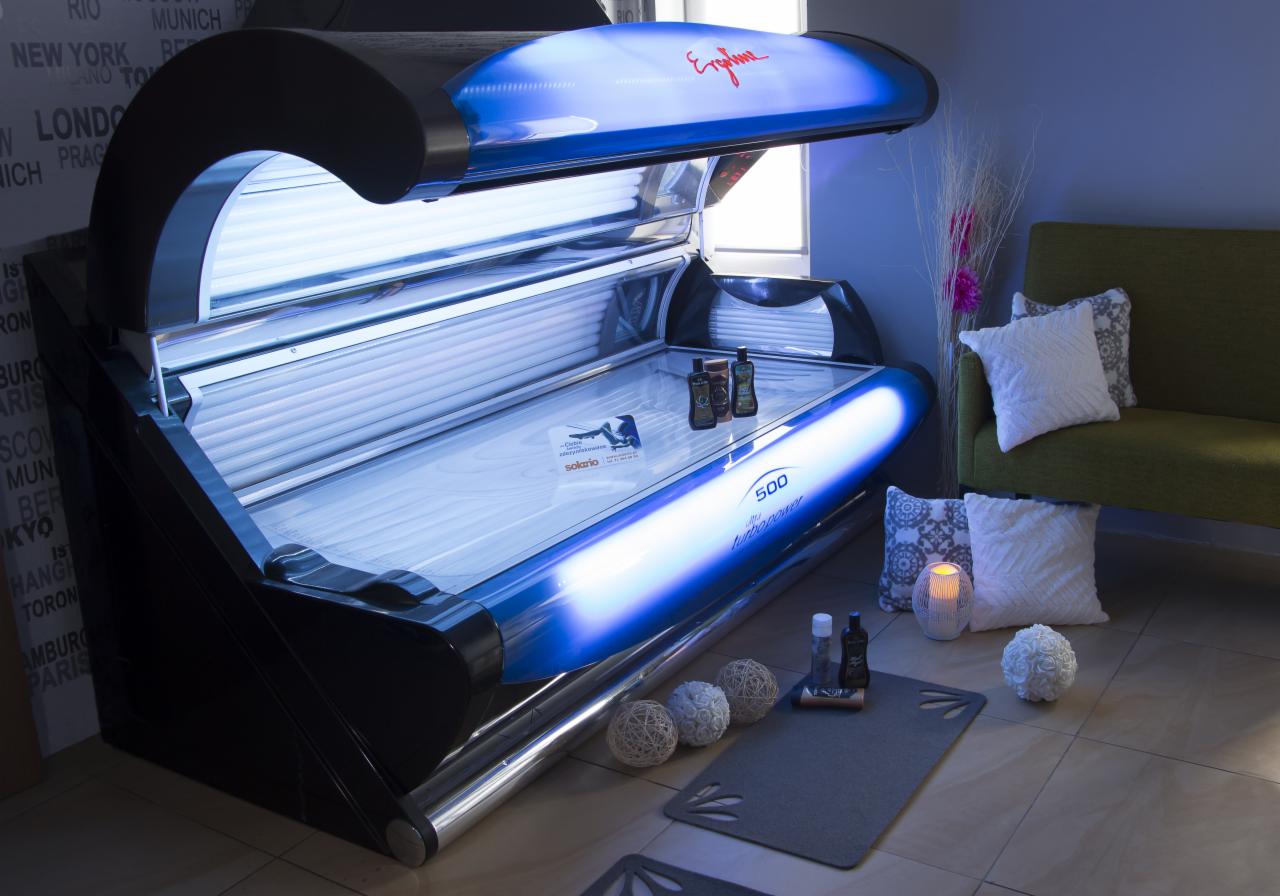 Otwarta kabina solarium Ergoline 500 z niebieskim światłem, kosmetyki do opalania na leżance i na podłodze, w tle sofa z poduszkami i dekoracje.