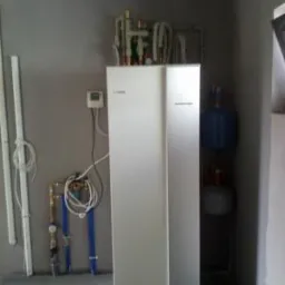 Biały piec grzewczy z podłączonymi rurami miedzianymi i niebieskimi, zamontowany na szarej podłodze w surowym pomieszczeniu z szarymi ścianami i widocznymi przewodami elektrycznymi.