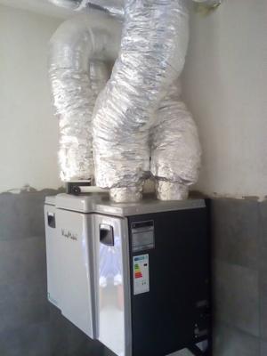 Szaro-czarna jednostka rekuperacyjna z trzema srebrnymi, elastycznymi przewodami wentylacyjnymi u góry, zamontowana przy ścianie z szarą lamperią.