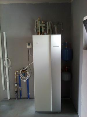 Biały piec grzewczy z podłączonymi rurami miedzianymi i niebieskimi, zamontowany na szarej podłodze w surowym pomieszczeniu z szarymi ścianami i widocznymi przewodami elektrycznymi.