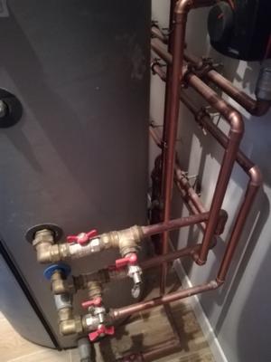 Szarej barwy zbiornik z podłączonymi miedzianymi rurami i zaworami z czerwonymi pokrętłami, widoczne złącza i fragment ściany w tle.