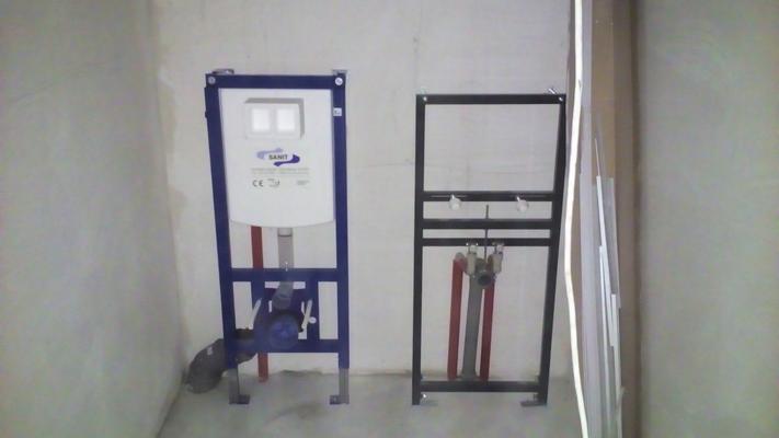 Stelaże podtynkowe do WC marki Sanit, jeden z niebieską ramą, drugi z czarną, z widocznymi rurami i przyłączami, na tle surowych ścian przed zabudową.