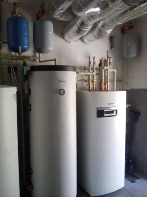 Dwa białe, pionowe zbiorniki buforowe Buderus w kotłowni, z widocznymi rurami miedzianymi i izolowanymi przewodami wentylacyjnymi pod sufitem.