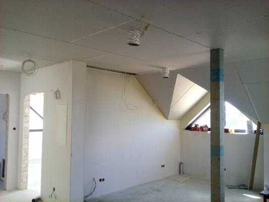 Poddasze w trakcie wykończenia: widoczne płyty gipsowo-kartonowe na suficie, instalacje elektryczne, pion konstrukcyjny zabezpieczony taśmą, okno dachowe.