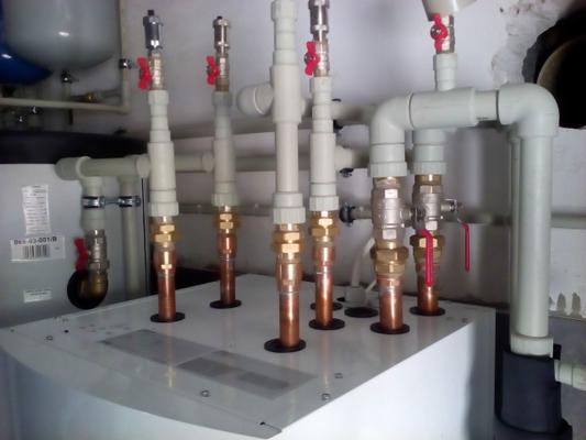 Nowoczesna instalacja centralnego ogrzewania z rur miedzianych i zaworów z czerwonymi uchwytami, widoczne połączenia z białymi rurami PCV, zamontowana na białym zbiorniku z czarną podstawą.