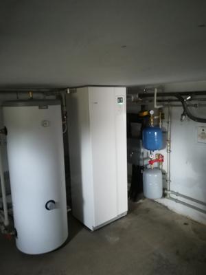 Nowoczesna, biała pompa ciepła w kotłowni, obok biały zasobnik ciepłej wody, niebieski i szary zbiornik wyrównawczy, widoczne rury instalacji, surowy betonowy sufit.