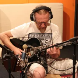 Mężczyzna z rudą brodą w słuchawkach gra na czarnej gitarze akustycznej w studiu nagrań, mikrofon pojemnościowy ustawiony blisko pudła rezonansowego.