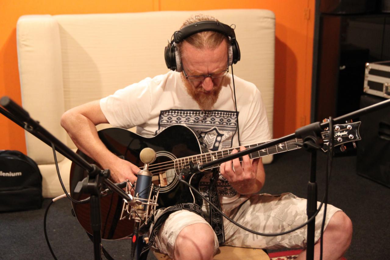 Mężczyzna z rudą brodą w słuchawkach gra na czarnej gitarze akustycznej w studiu nagrań, mikrofon pojemnościowy ustawiony blisko pudła rezonansowego.