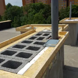 Konstrukcja kominowa na dachu płaskim w trakcie budowy, widoczne bloczki betonowe, drewniana rama i pionowa rura wentylacyjna na tle bloków mieszkalnych.