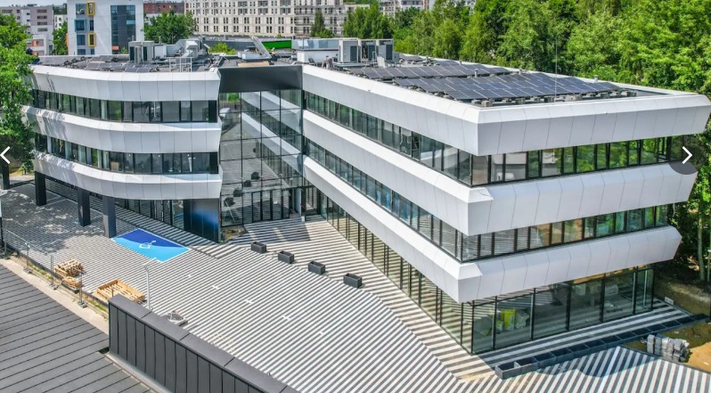 Nowoczesny, trzypiętrowy budynek biurowy z panelami słonecznymi na dachu, widoczny z zewnątrz, z charakterystyczną elewacją i wzorem na chodniku.