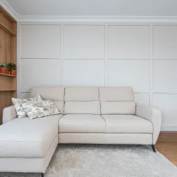 Jasny salon z beżową sofą narożną, dekoracyjną ścianą w panelach i drewnianym regałem z zegarem. Stonowana kolorystyka, minimalistyczny design.
