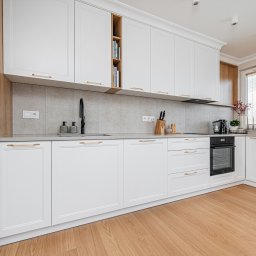 KartA Pracownia Projektowa Katarzyna Cimochowska - Biała kuchnia z drewnianymi akcentami. Fronty szafek z frezowaniami, złote uchwyty. Na blacie czarny zlew i bateria. Na półkach książki i dekoracje.