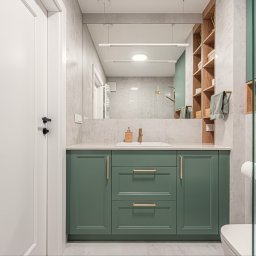 KartA Pracownia Projektowa Katarzyna Cimochowska - Nowoczesna łazienka z zielonymi szafkami pod umywalką, dużym lustrem i drewnianymi półkami. Minimalistyczny design, jasne płytki, złote akcenty.