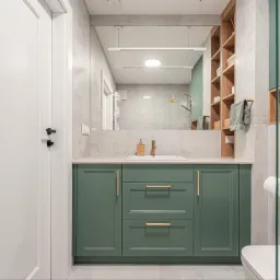 Nowoczesna łazienka z zielonymi szafkami pod umywalką, dużym lustrem i drewnianymi półkami. Minimalistyczny design, jasne płytki, złote akcenty.