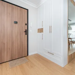 Jasne wnętrze przedpokoju: drewniane drzwi z czarną ramą, biała szafa z złotymi uchwytami, lustro odbijające jadalnię, drewniana podłoga i wycieraczka.