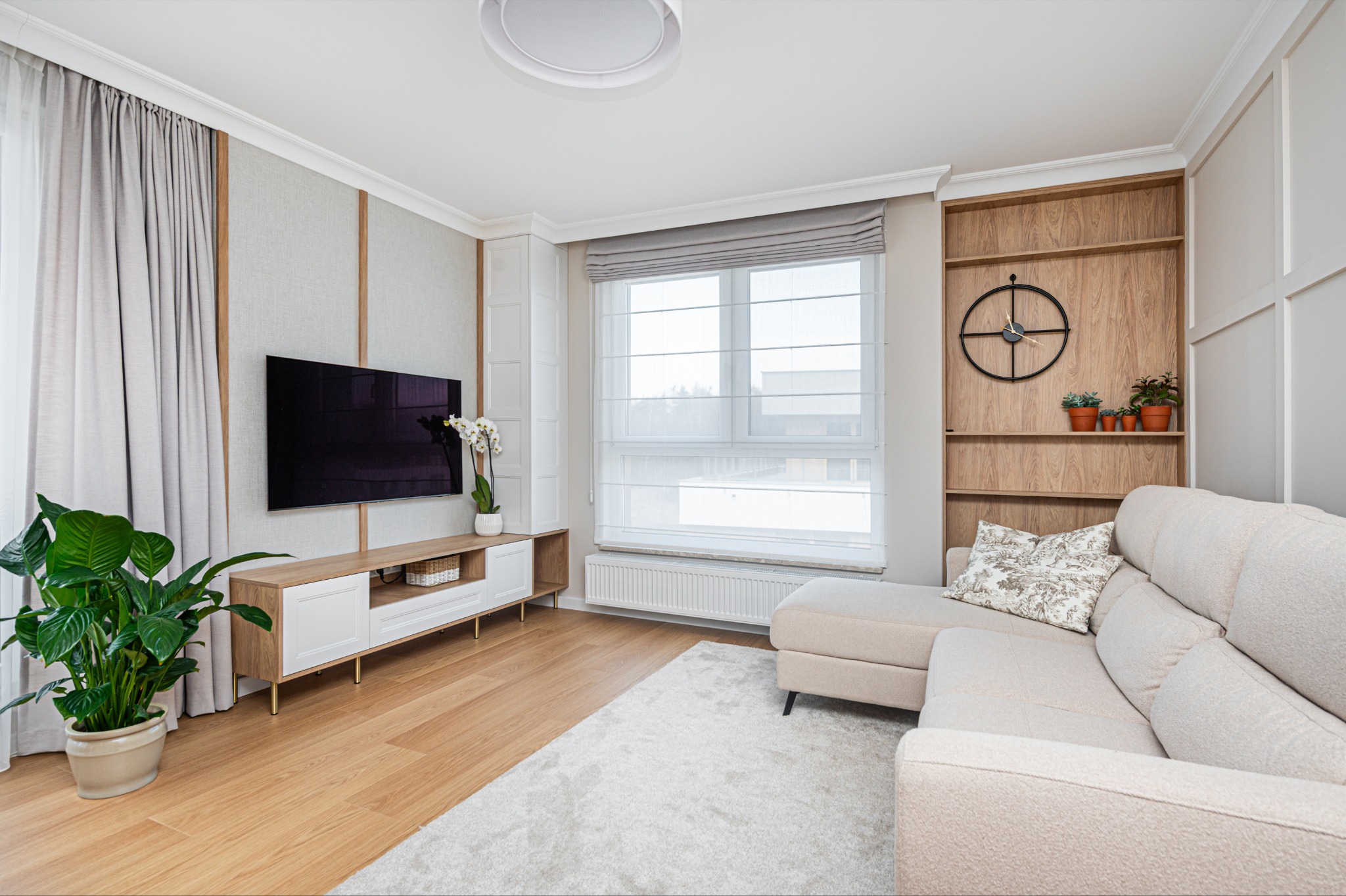 Jasne wnętrze salonu z beżową sofą, telewizorem na ścianie i drewnianymi akcentami. Roślina doniczkowa dodaje świeżości. Nowoczesny design z dbałością o detale.