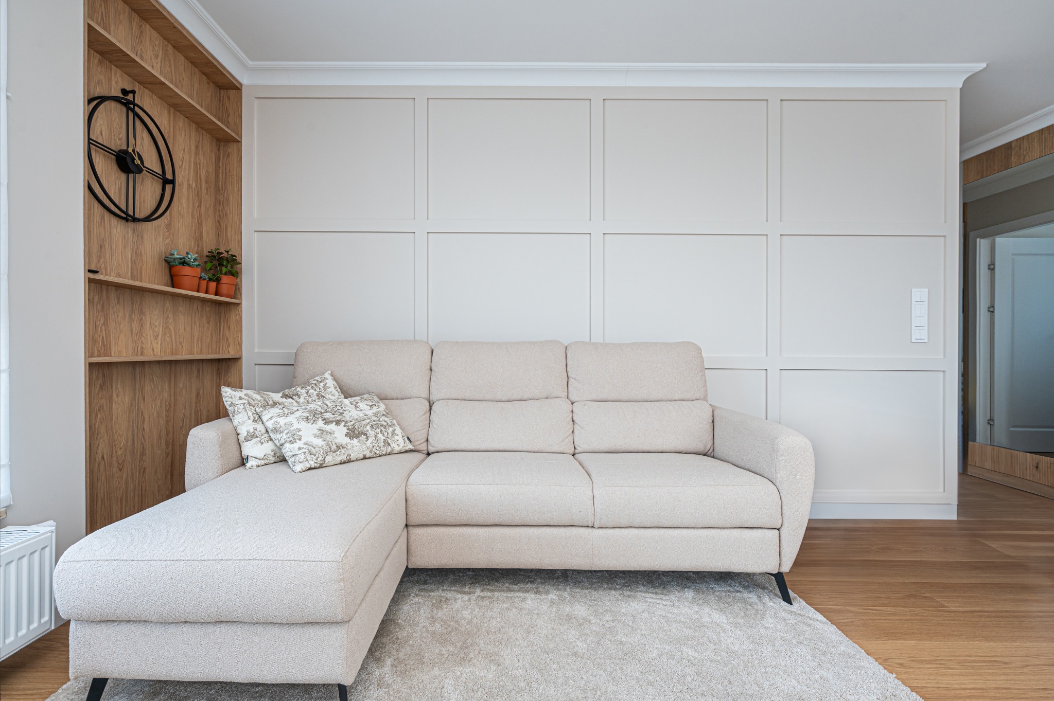 Jasny salon z beżową sofą narożną, dekoracyjną ścianą w panelach i drewnianym regałem z zegarem. Stonowana kolorystyka, minimalistyczny design.