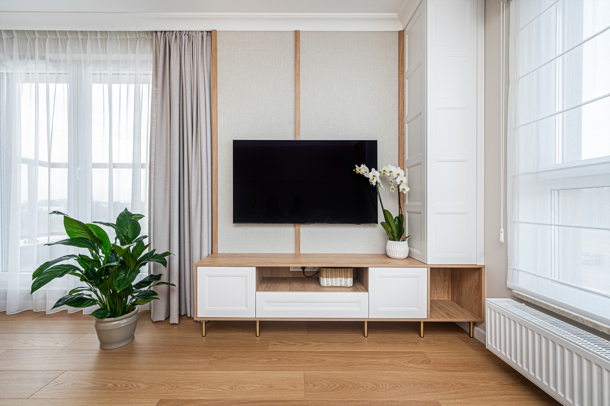 Nowoczesny salon z drewnianą podłogą, beżowymi zasłonami i ścianą TV z szafką RTV w odcieniach bieli i drewna. Na szafce orchidea w doniczce.