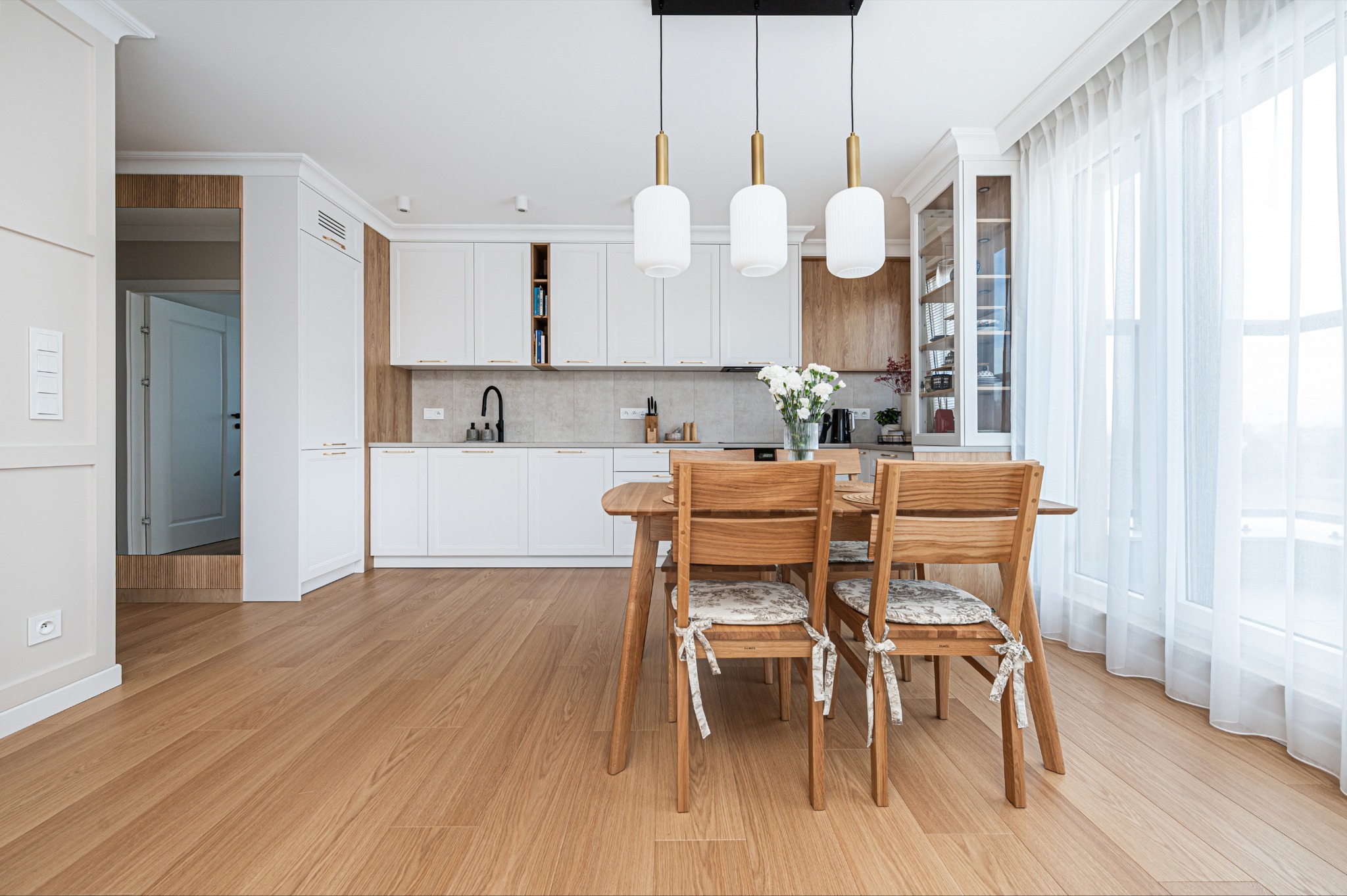Jasna, nowoczesna kuchnia z drewnianym stołem i krzesłami. Białe szafki z drewnianymi akcentami, trzy wiszące lampy i bukiet białych kwiatów na stole.