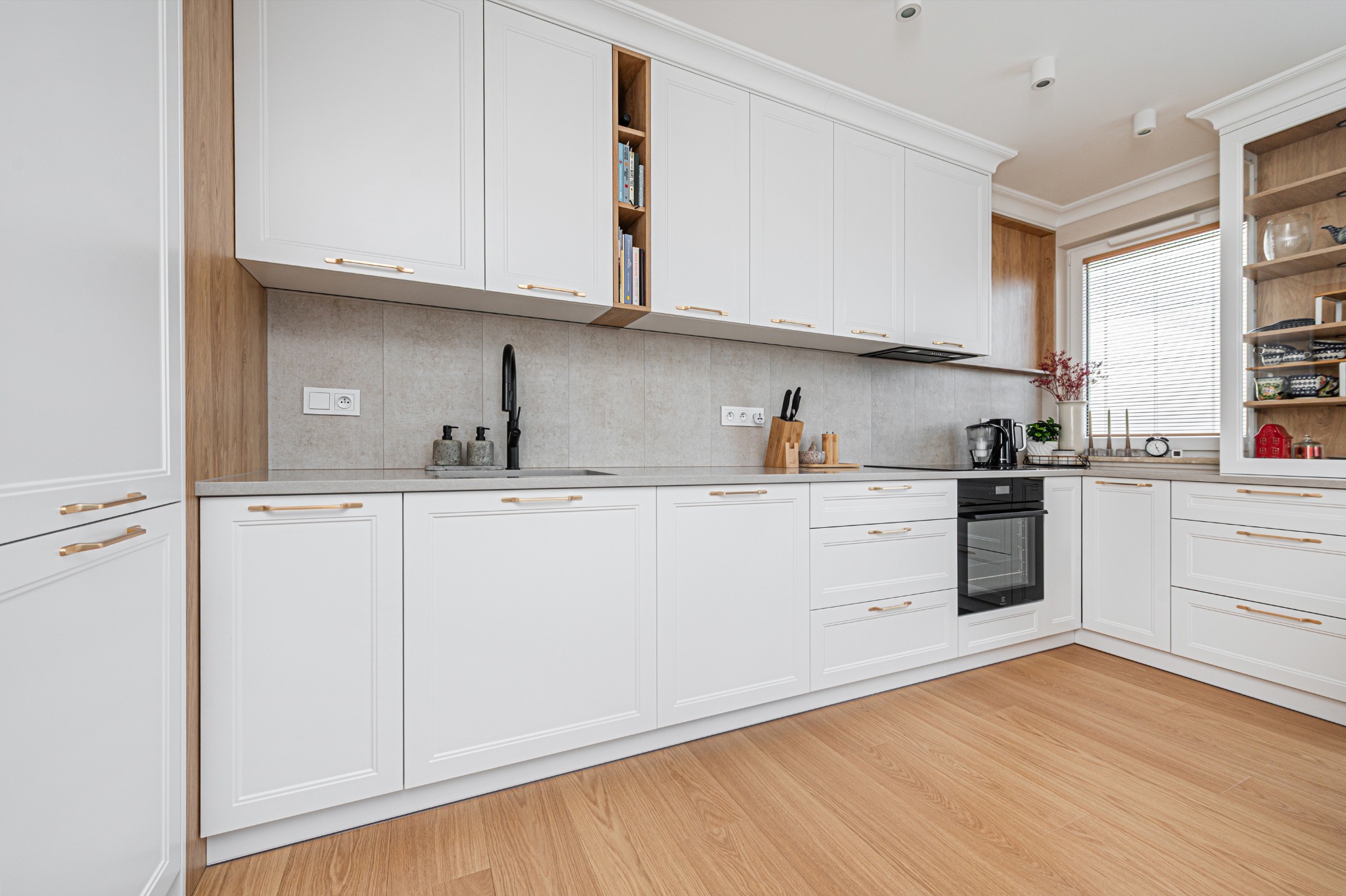 Biała kuchnia z drewnianymi akcentami. Fronty szafek z frezowaniami, złote uchwyty. Na blacie czarny zlew i bateria. Na półkach książki i dekoracje.