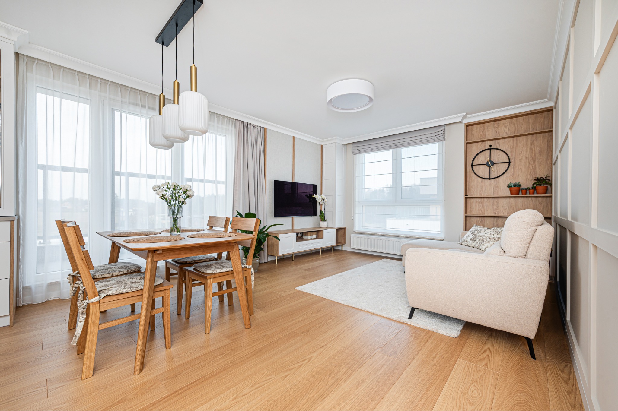 Jasne, przestronne wnętrze: drewniany stół z kwiatami, krzesła z poduszkami, sofa na dywanie, TV na komodzie, okna z zasłonami i lampy wiszące.