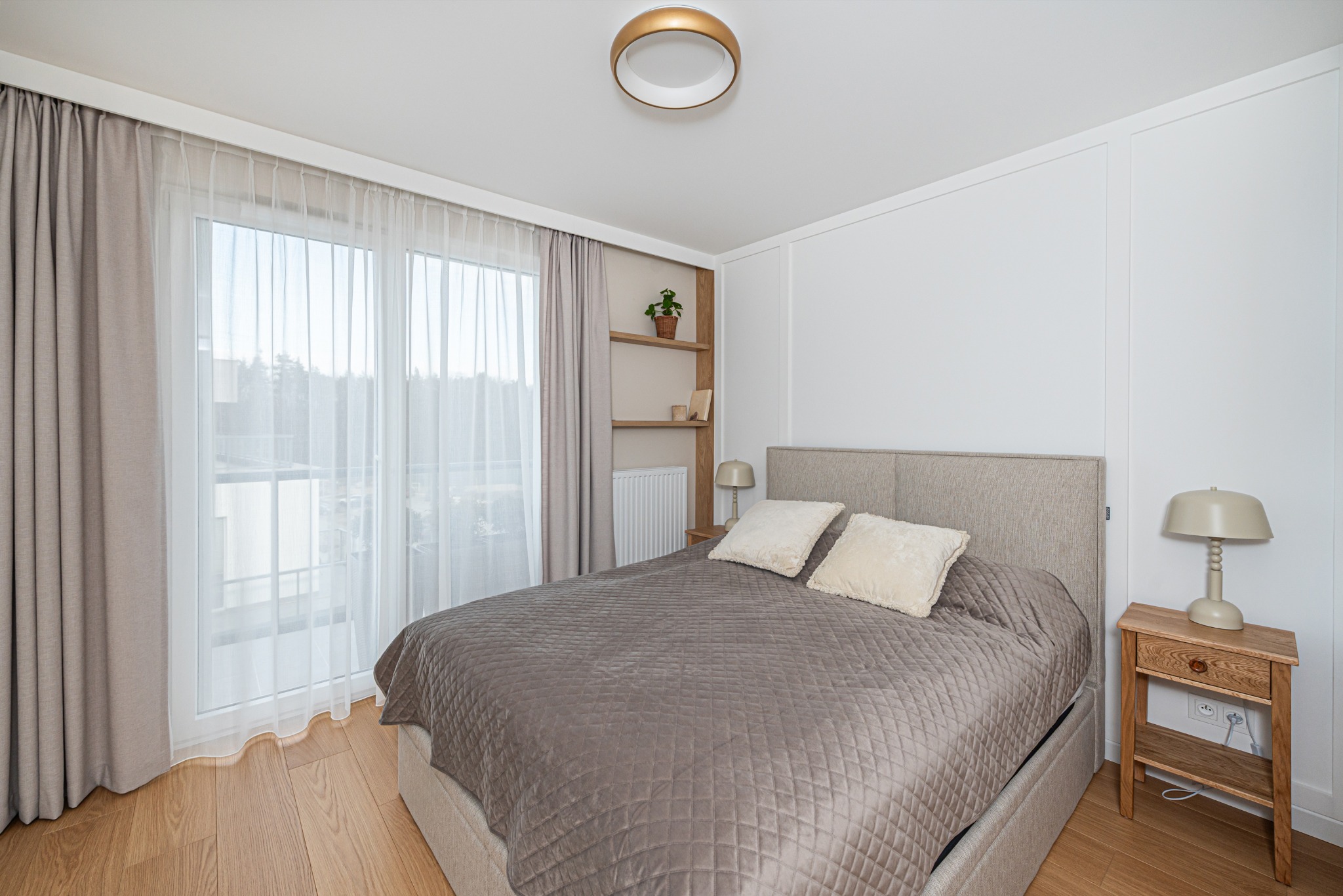 Jasna sypialnia z drewnianą podłogą i beżowymi zasłonami. Na ścianie półki z dekoracjami i roślinką.  Na szafce nocnej stoi lampka.