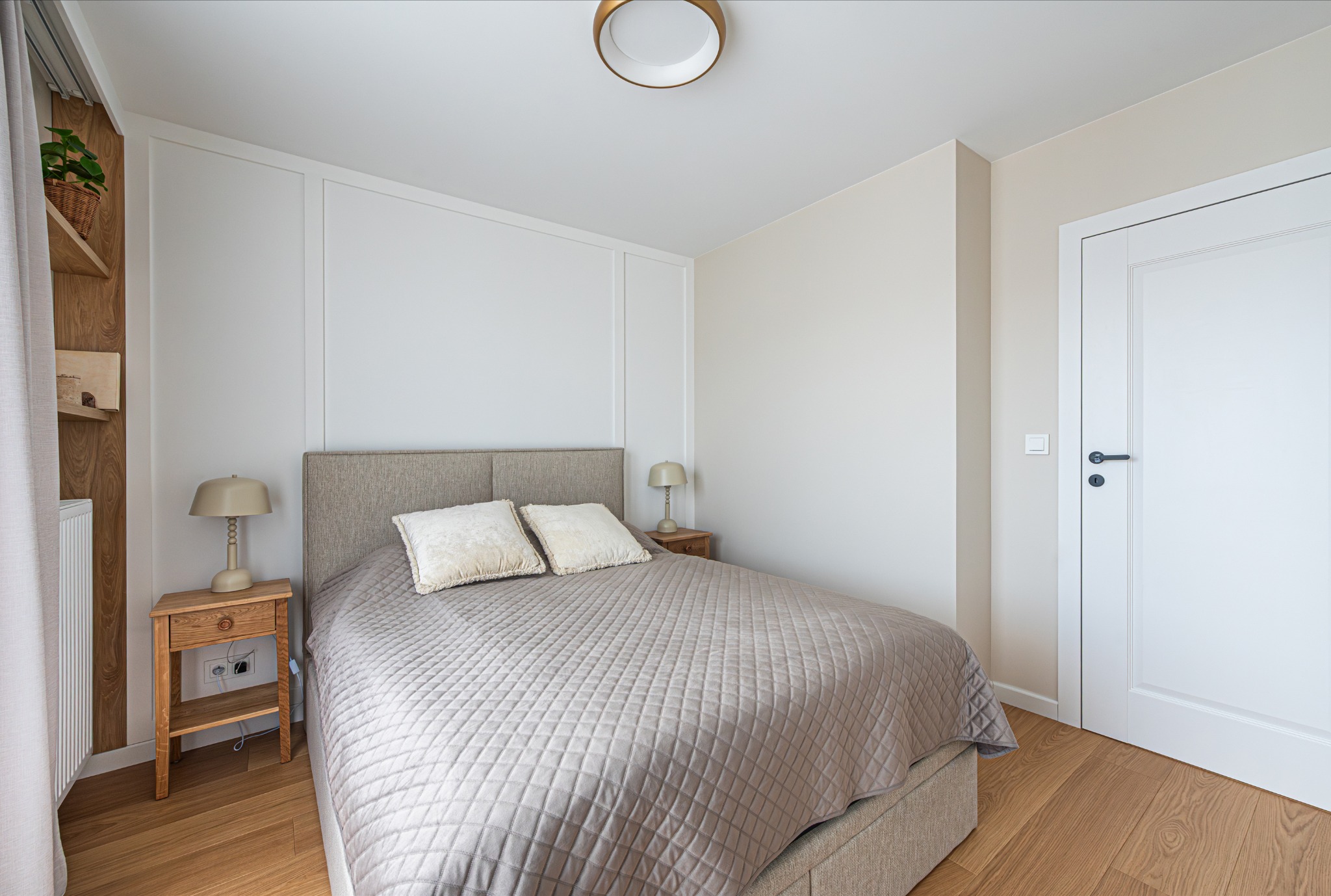 Neutralna sypialnia z drewnianą podłogą, beżowym łóżkiem i białymi ścianami z dekoracyjnymi panelami. Dwie szafki nocne z lampkami i półki z rośliną.