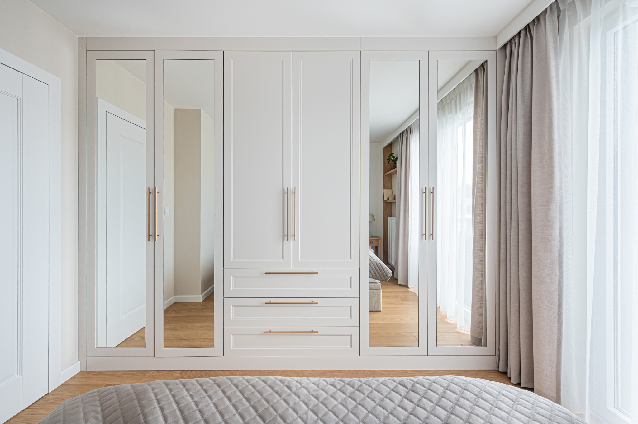 Biała szafa wnękowa z lustrami i złotymi uchwytami w jasnej sypialni. Widoczne odbicie wnętrza pokoju i fragment łóżka z pikowaną narzutą.