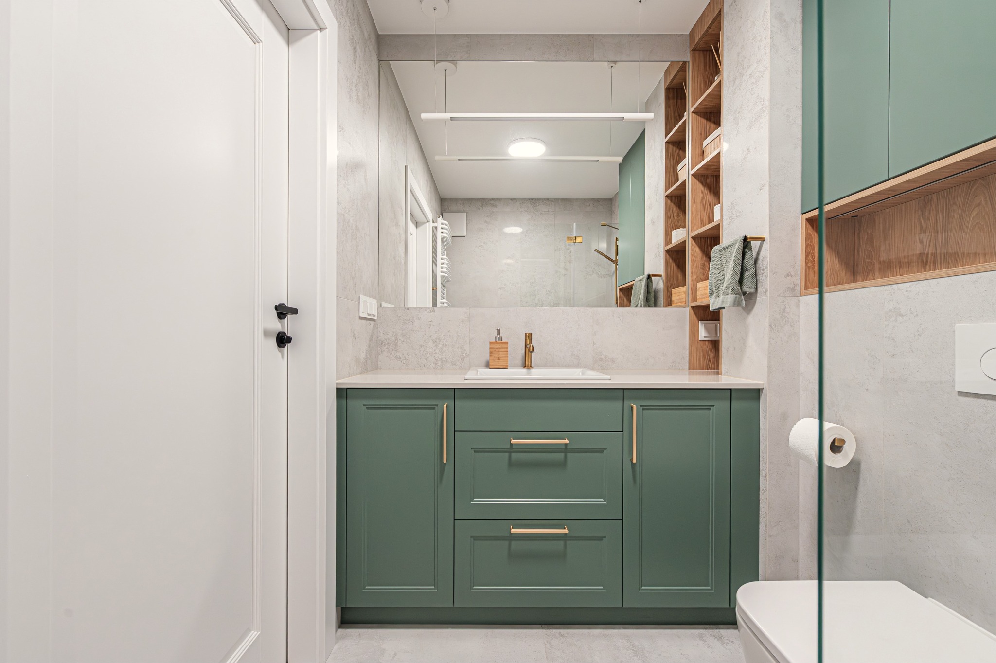 Nowoczesna łazienka z zielonymi szafkami pod umywalką, dużym lustrem i drewnianymi półkami. Minimalistyczny design, jasne płytki, złote akcenty.