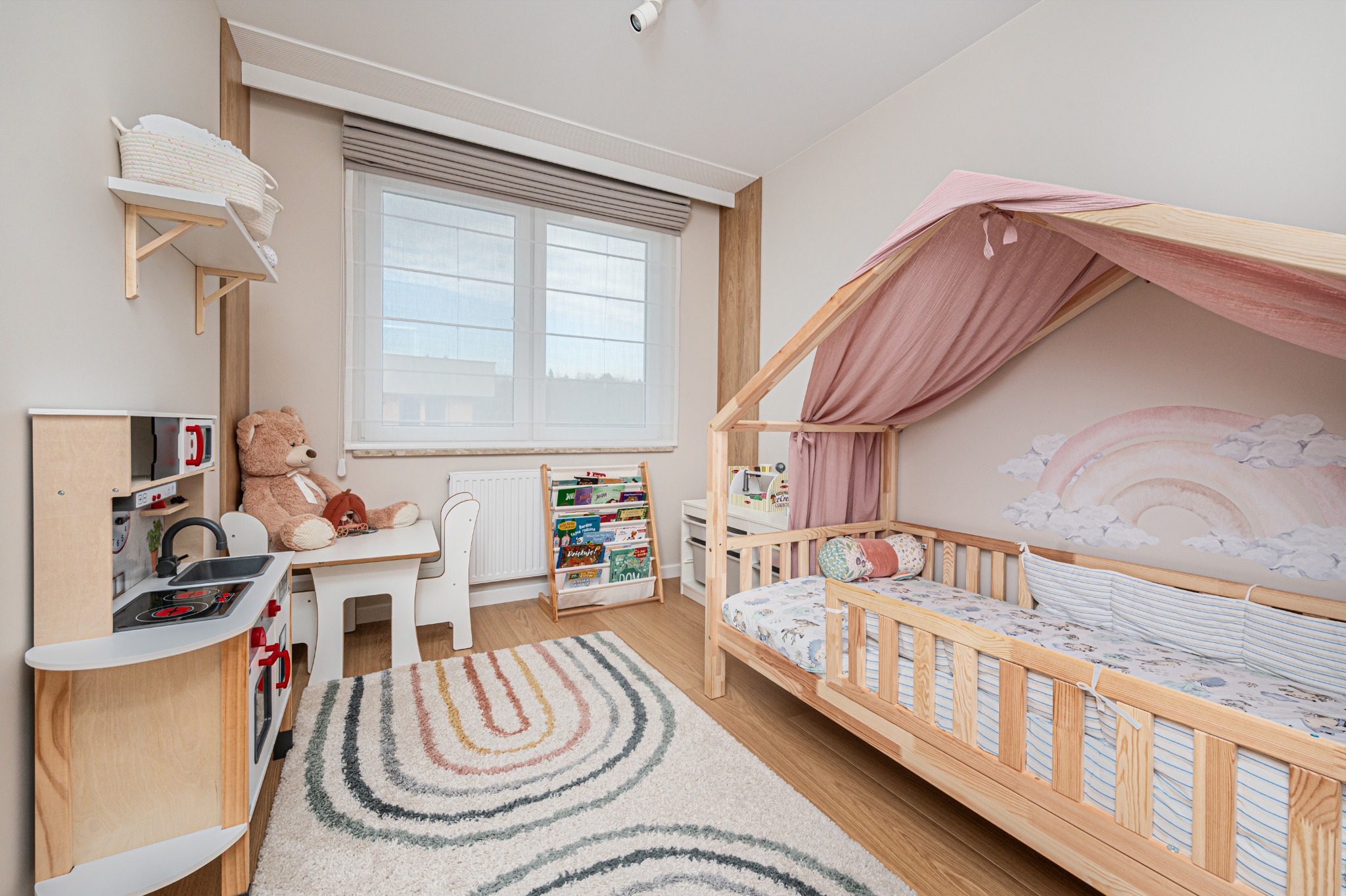 Pokój dziecięcy w pastelowych barwach: drewniane łóżko z baldachimem, dywan w tęczę, zabawki i regał z książkami tworzą przytulną przestrzeń.