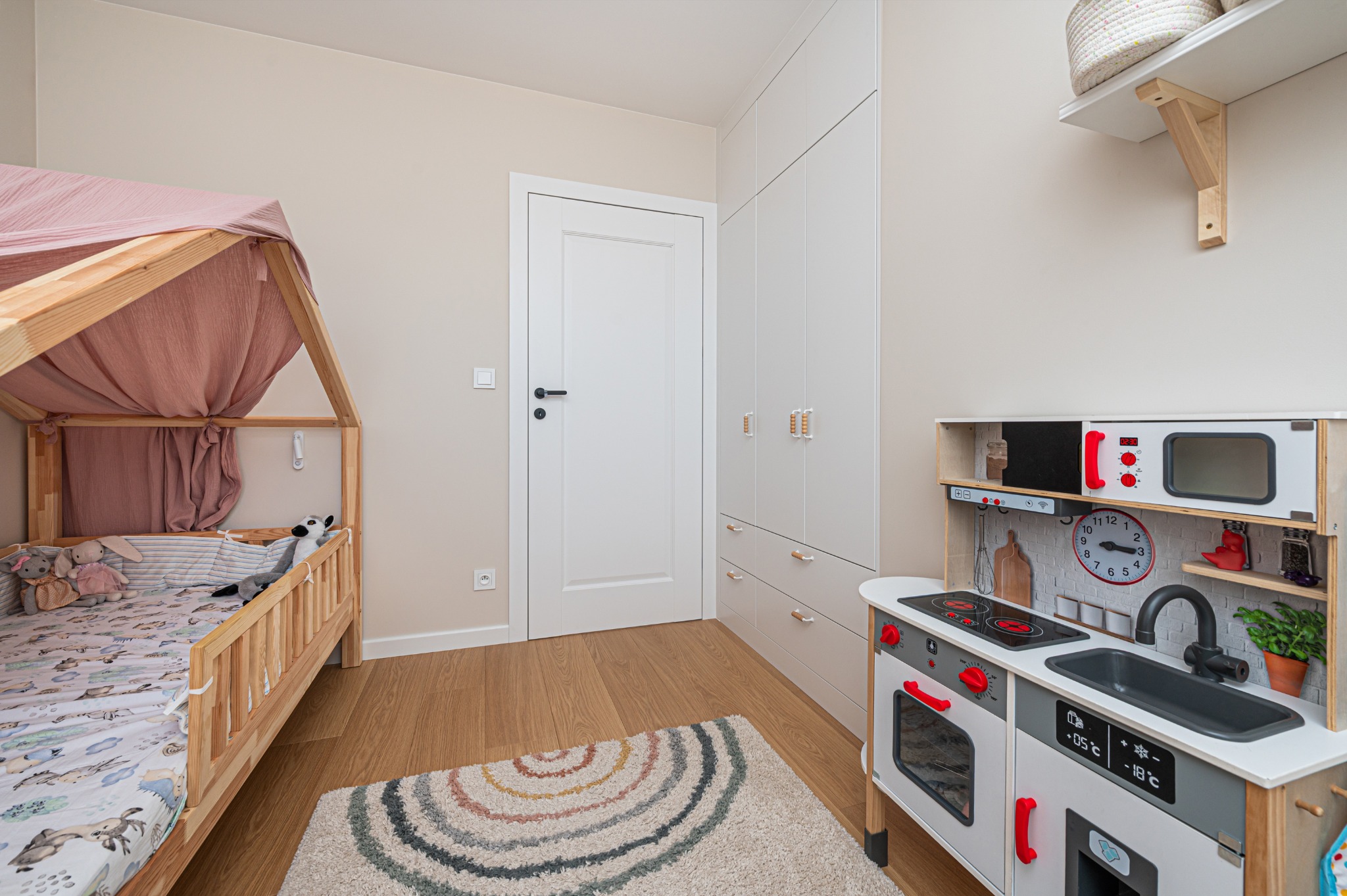 Pokój dziecka w neutralnych barwach: drewniane łóżko z baldachimem, biała szafa, drewniana kuchnia zabawkowa i okrągły dywan na drewnianej podłodze.