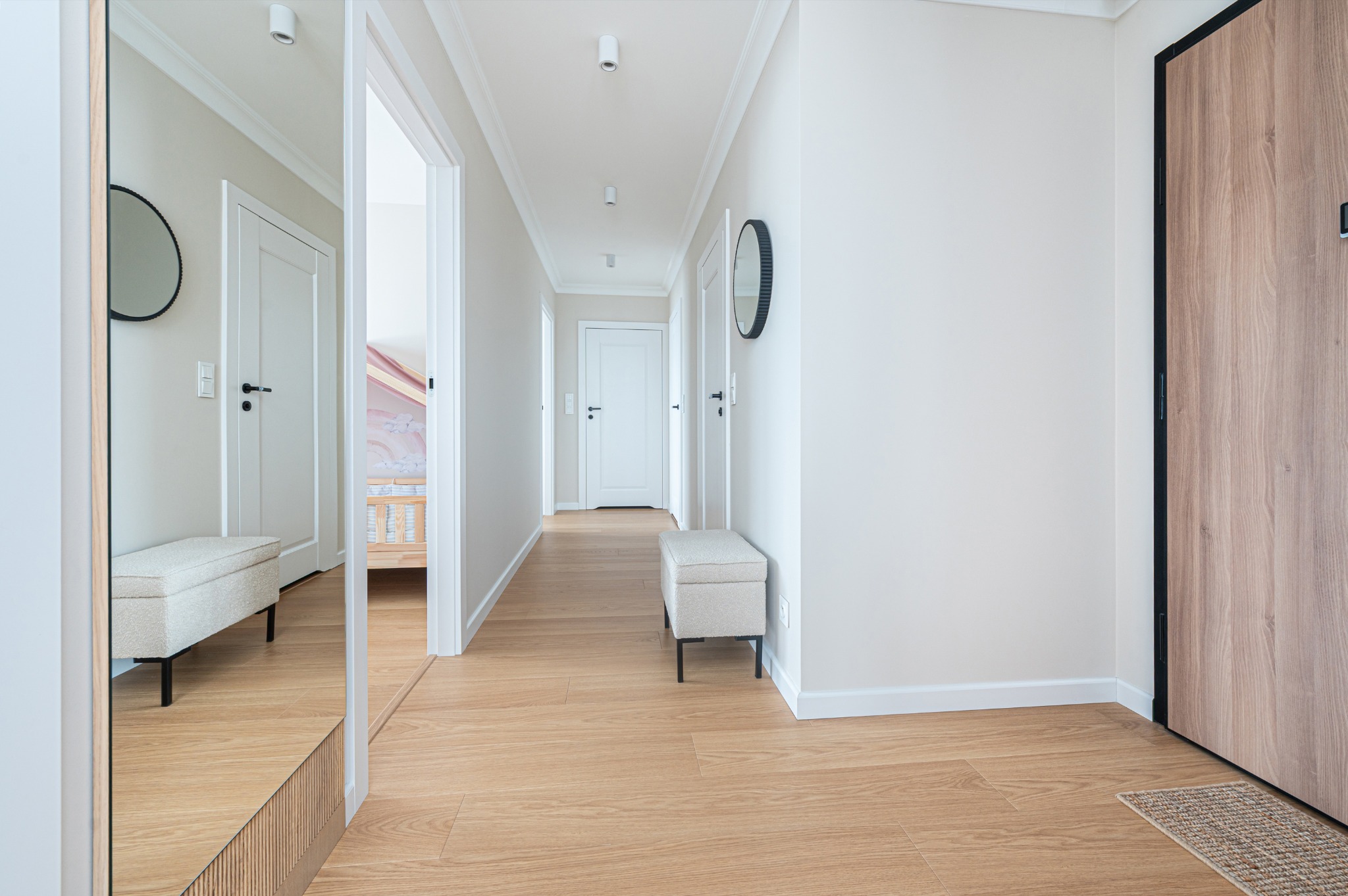 Jasny korytarz z drewnianą podłogą, lustrem i pufą. Widoczne białe drzwi i okrągłe lustra na ścianach. Minimalistyczny design.