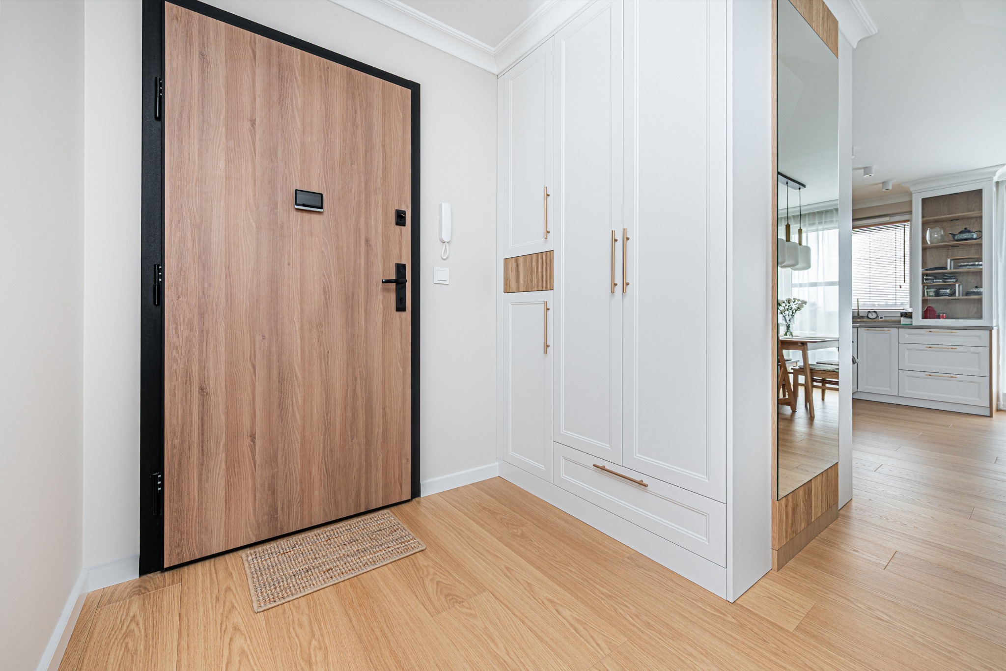 Jasne wnętrze przedpokoju: drewniane drzwi z czarną ramą, biała szafa z złotymi uchwytami, lustro odbijające jadalnię, drewniana podłoga i wycieraczka.