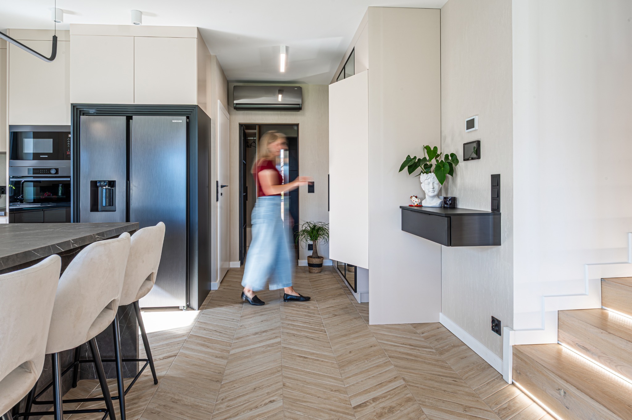 Nowoczesne wnętrze: parkiet w jodełkę, minimalistyczna konsola z dekoracjami, schody z podświetleniem LED, kobieta w tle. Jasna kolorystyka i przemyślany design.