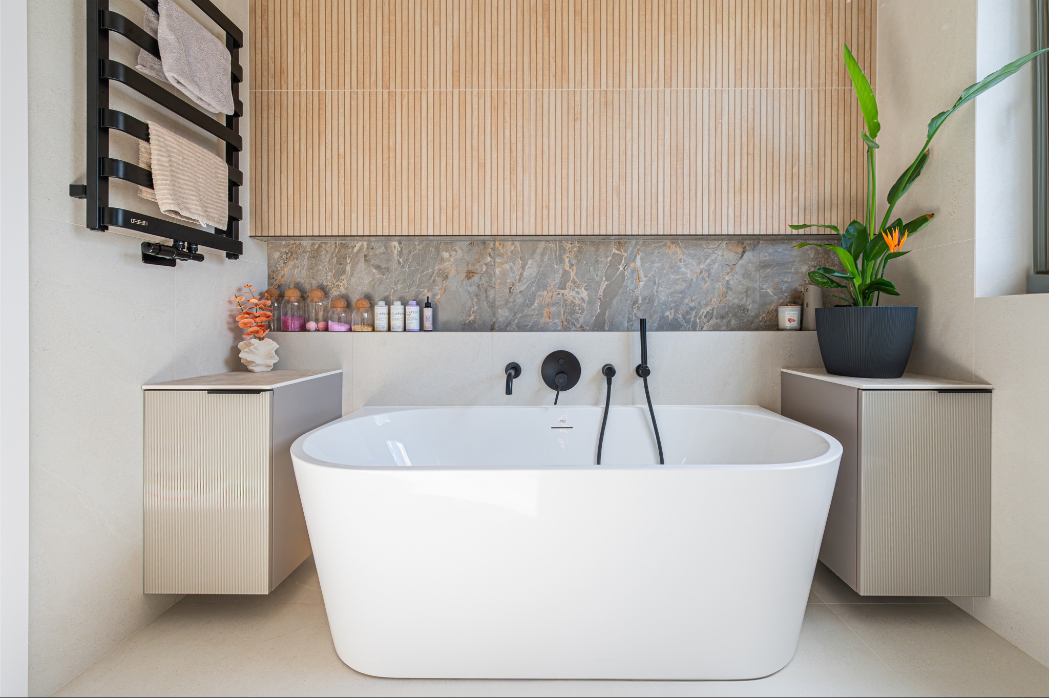 Nowoczesna łazienka z wolnostojącą białą wanną, armaturą w kolorze czarnym, drewnianą ścianą i szafkami w odcieniach beżu. Minimalistyczny design z rośliną doniczkową.