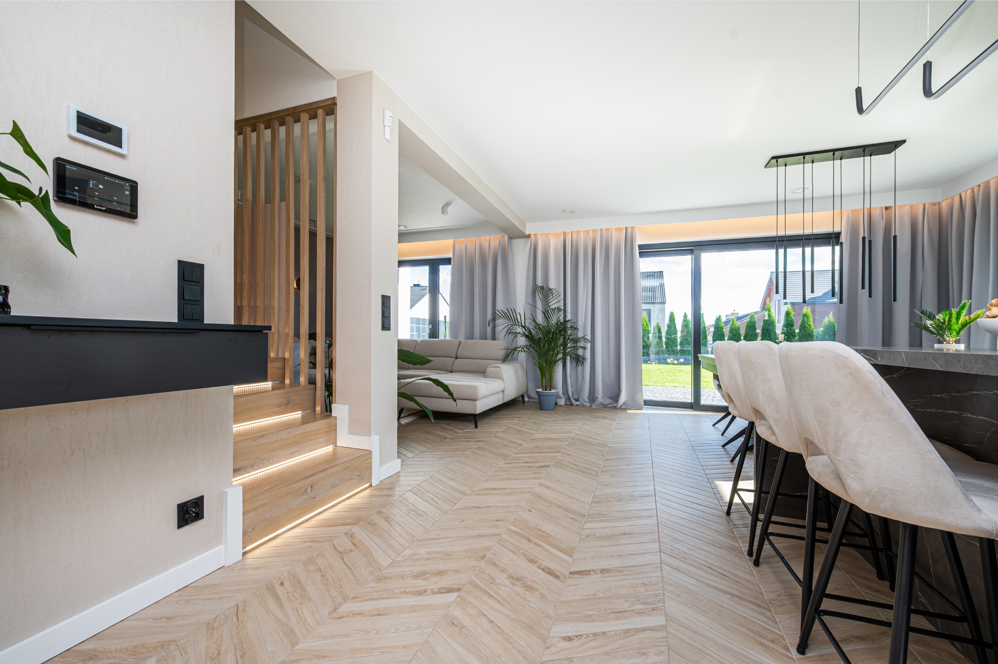 Jasne, przestronne wnętrze domu z drewnianą podłogą w jodełkę, schodami z podświetleniem LED i widokiem na ogród przez duże okna. Nowoczesny design z elementami naturalnymi.