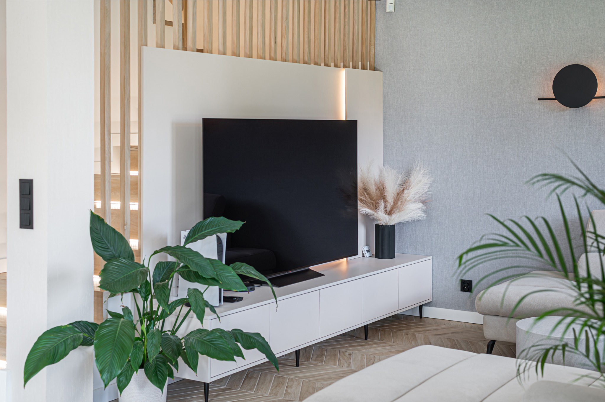 Nowoczesny salon z telewizorem, białą szafką RTV, roślinami doniczkowymi i drewnianymi lamelami w tle. Minimalistyczny design z akcentami naturalnymi.