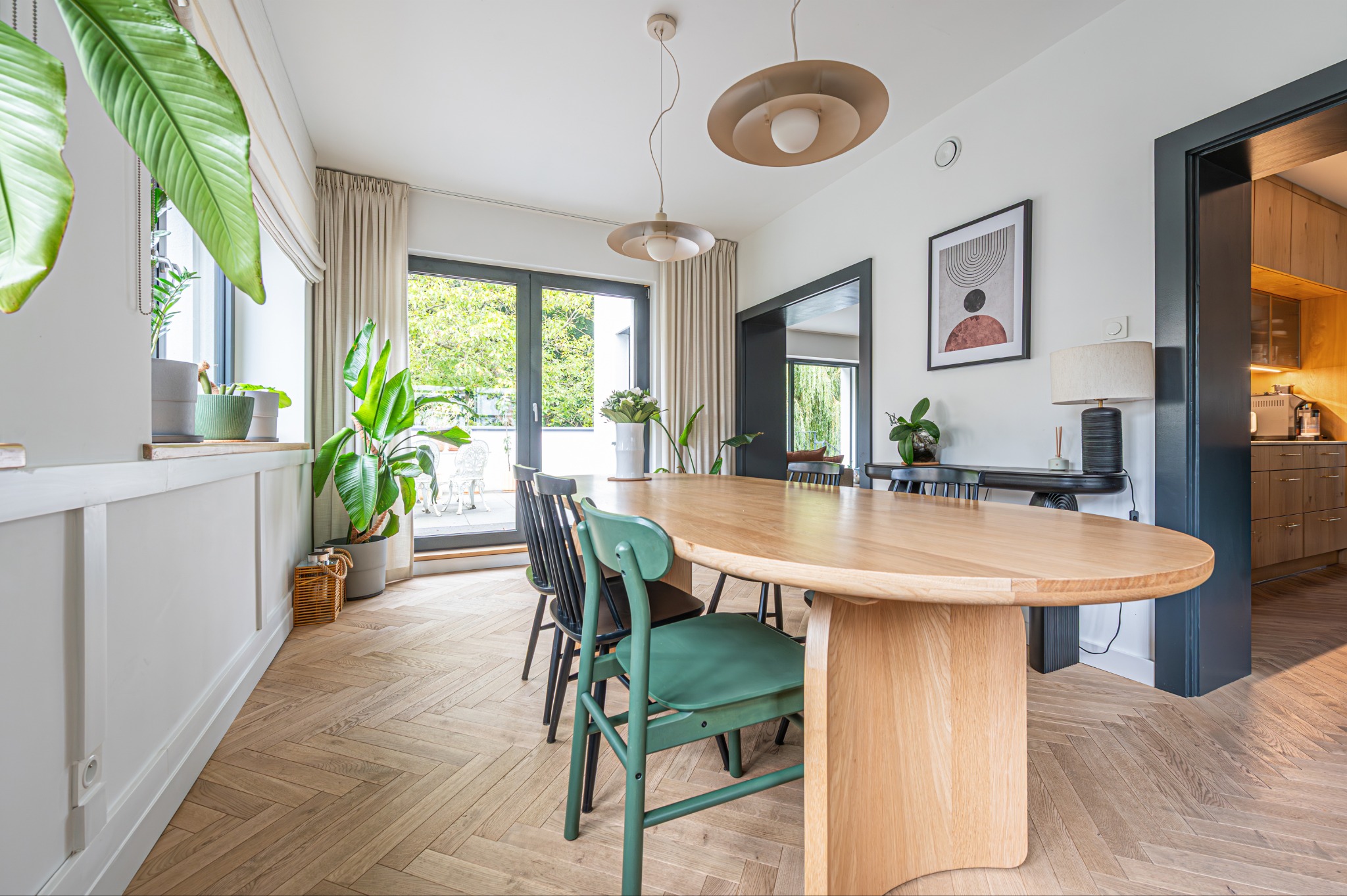Jasna jadalnia z drewnianym stołem i parkietem w jodełkę. Widok na balkon przez okno. Minimalistyczne lampy i dekoracje. W tle otwarta kuchnia.