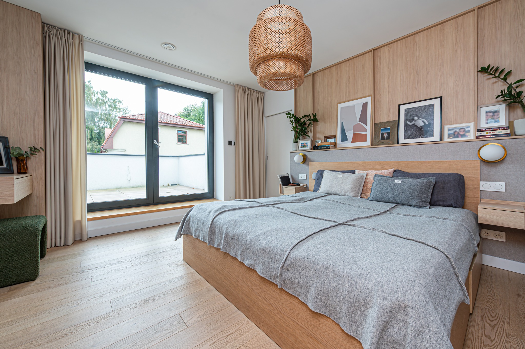 Jasna sypialnia z drewnianym wykończeniem, dużym oknem balkonowym i plecioną lampą. Neutralna kolorystyka, minimalistyczny design, widok na zewnątrz.