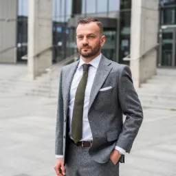 Mężczyzna w szarym garniturze i zielonym krawacie stoi przed nowoczesnym budynkiem biurowym z przeszkloną fasadą.