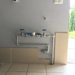 Fragment instalacji wodnej z zaworami i licznikiem, zamontowanej na szarej ścianie nad beżowymi płytkami podłogowymi i ściennymi, widoczne elementy instalacji z tworzywa i metalu.