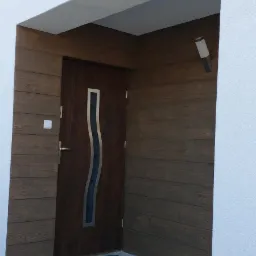 Wejście do domu z elewacją imitującą drewno, brązowe drzwi z przeszkleniem o falistym kształcie, betonowe schody wejściowe i nowoczesna lampa ścienna.