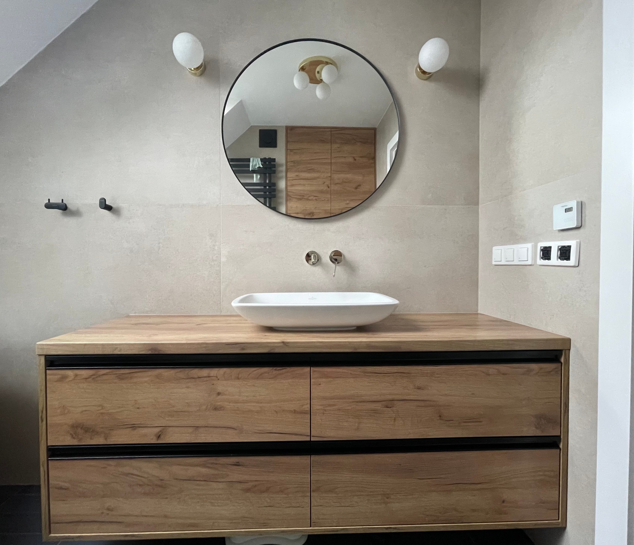 Łazienka w beżowych tonacjach z drewnianą szafką pod umywalkę z czterema szufladami z czarnymi wstawkami, okrągłe lustro odbija wnętrze z drewnianą szafą, nad szafką biała umywalka nablatowa,...