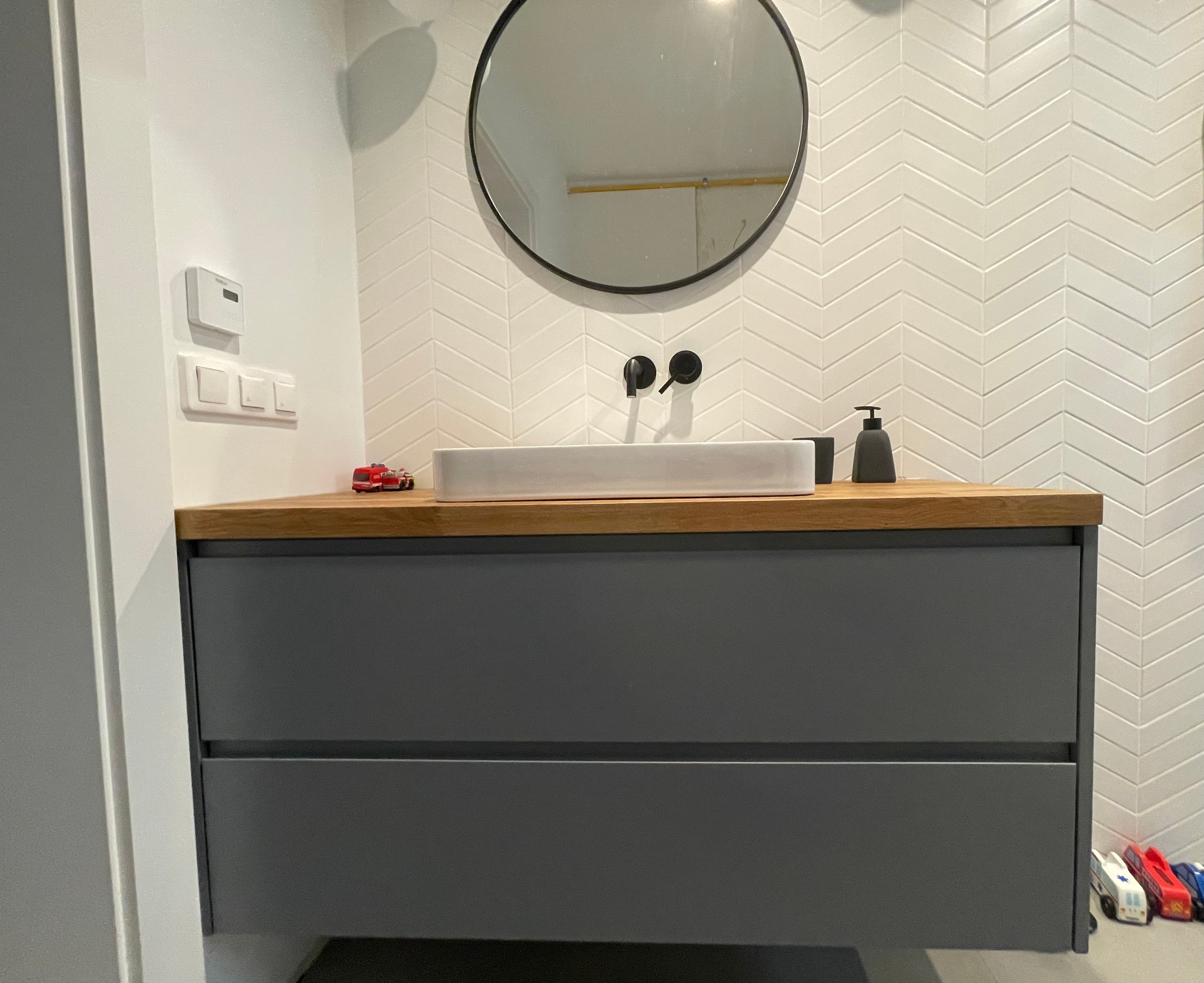 Szara szafka łazienkowa z drewnianym blatem, białą umywalką i czarną armaturą na tle białych płytek ułożonych w jodełkę; na blacie zabawkowy wóz strażacki, obok dozownik mydła, na podłodze zabawki.