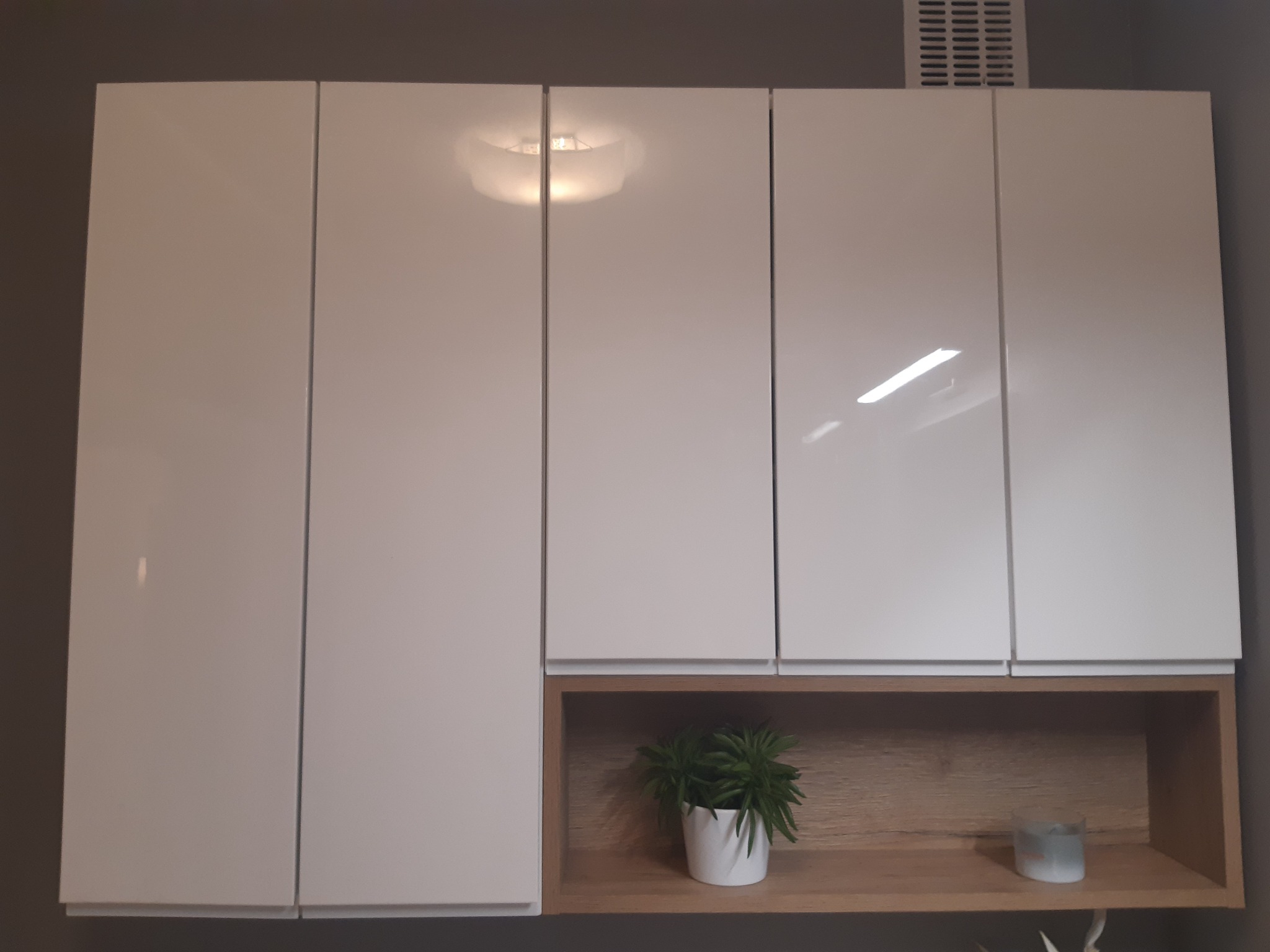 Białe, lakierowane szafki wiszące z otwartą półką z drewnianym wykończeniem, na której znajduje się doniczka z rośliną i świeczka.