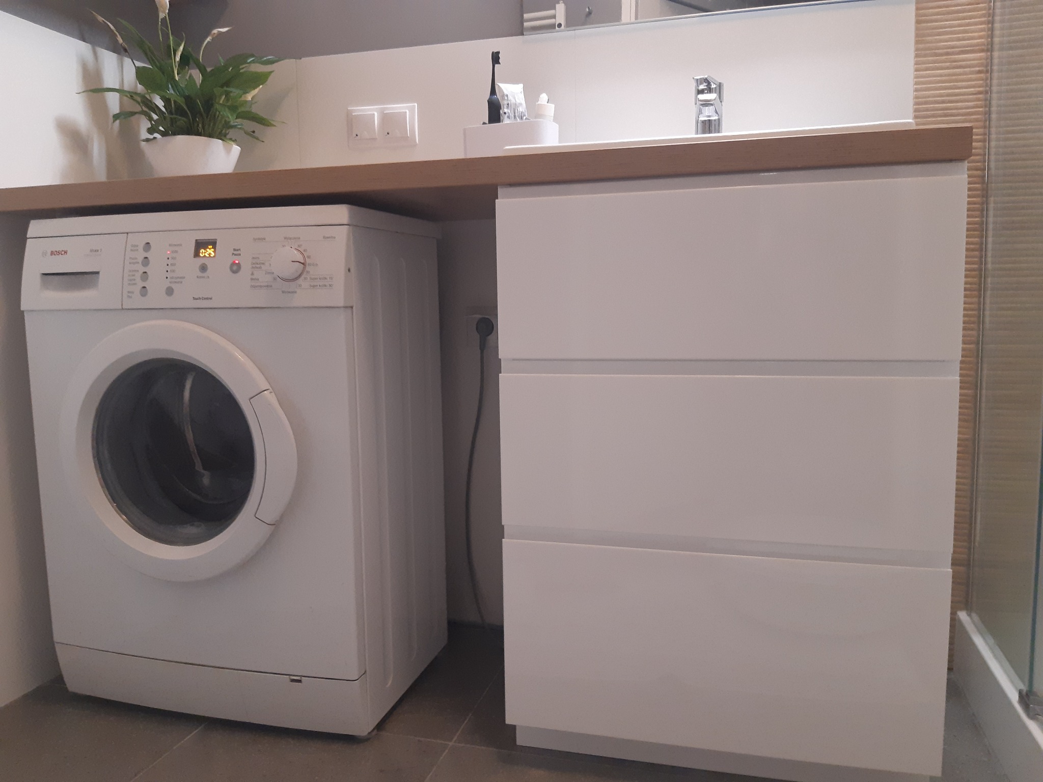 Biała pralka marki Bosch obok białej szafki z trzema szufladami pod drewnianym blatem w łazience, na blacie doniczka z zieloną rośliną.