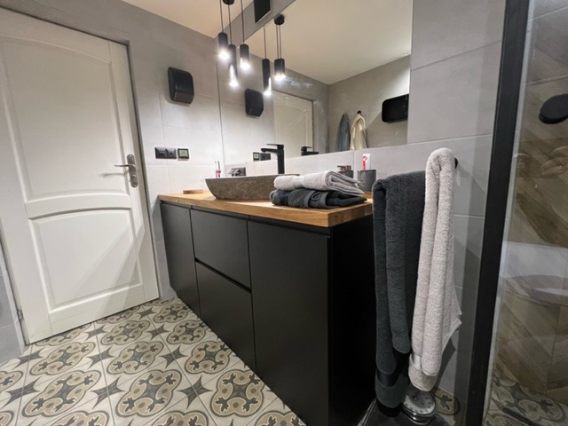 Nowoczesna łazienka z czarną szafką pod umywalkę z blatem drewnianym, kamienną umywalką, ozdobnymi płytkami podłogowymi i oświetleniem punktowym.