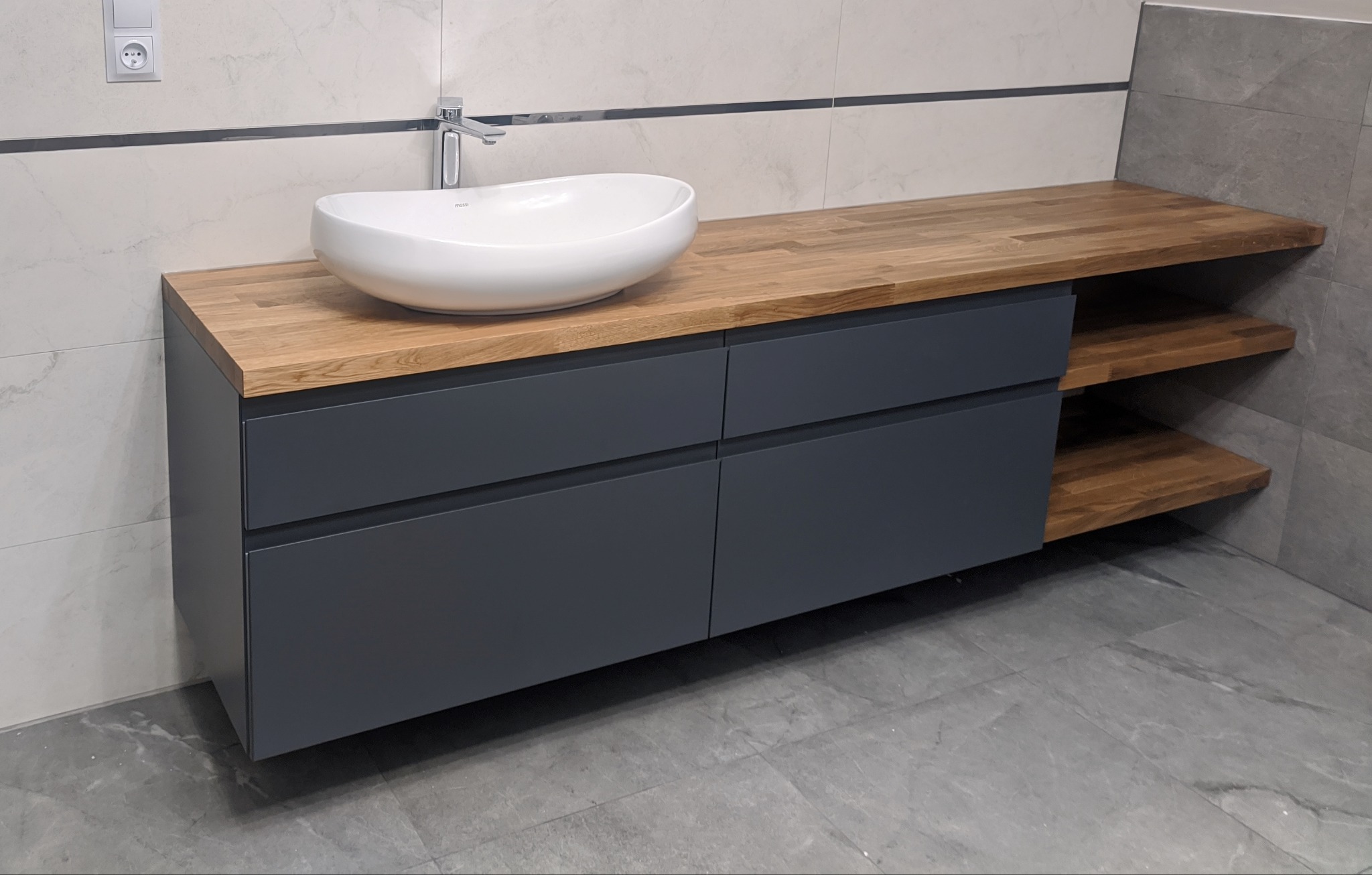 Szara szafka łazienkowa z drewnianym blatem i umywalką nablatową, uzupełniona o drewniane półki po prawej stronie, na tle marmurowych płytek.